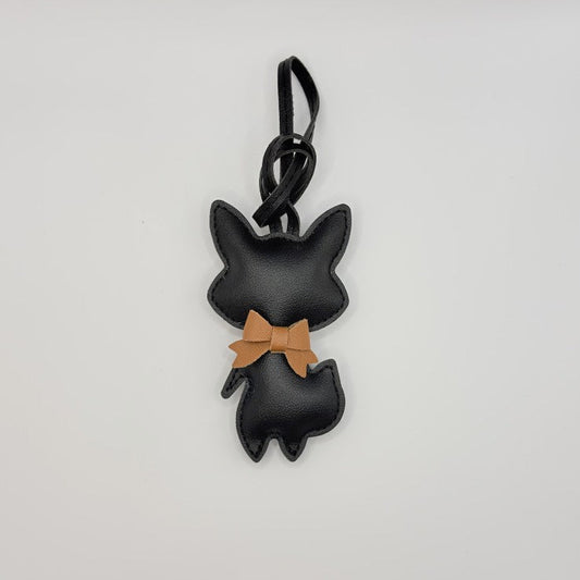 A black faux-leather keychain charm shaped like a cat silhouette with a tan bow tie, attached to a black strap, shown on a plain white background.